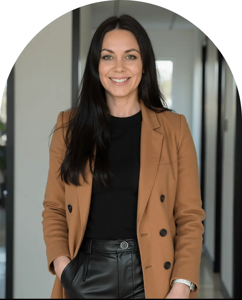 A woman wearing a black shirt and a brown blazer, standing confidently with a neutral expression.